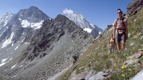 Zwei Wanderer auf dem Alpenpässeweg im Wallis mit Blick auf die steinige Berglandschaft im Hintergrund.