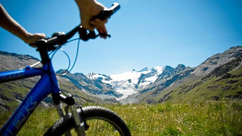 Der Lenker eines blauen Mountainbikes auf einer saftig grünen Wiese zwischen den Bergen bei strahlendem Sonnenschein.