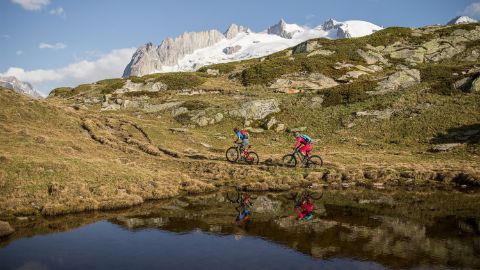 Zwei Mountaikbiker fahren über eine Wiese, hinter einem kleinen See und wundervollen, leicht mit Schnee bedekten Bergspitzen.