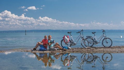 Des cyclistes lisent un magazine assis sur du gravier au bord du lac. La route du cœur. Vacances à vélo avec Eurotrek.