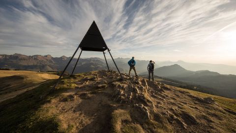 Two hikers stand on a mountain peak and gaze at the mountain landscape in front of them. Next to the hikers is a metal triangle with its tip pointing towards the sky.