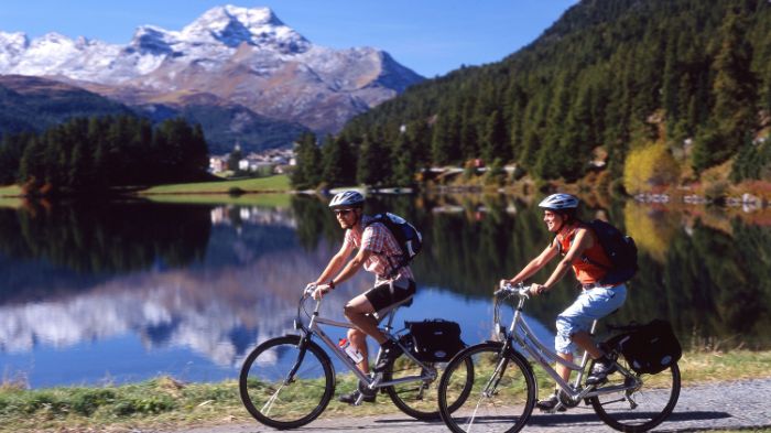 2 Biker fahren am Ufer des Silvaplanersees. im Hintergrund die mit Schneebedeckte Bergspitzen, welche sich auch im See spiegeln.