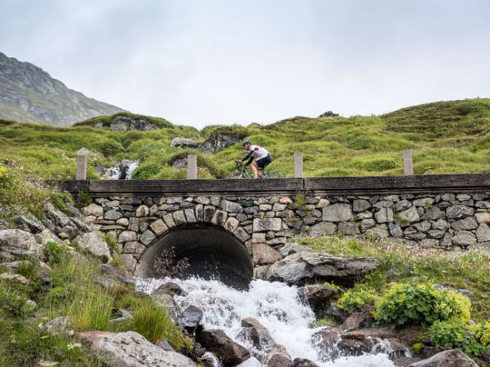 Ein Biker fährt über eine steinerne Brücke bei der aus einem Bogen das Wasser durchfliesst.