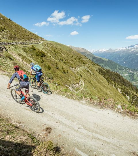 Zwei Mountainbiker auf einer Naturstrasse in den Bergen. Rechts eine Berglandschaft mit strahlend blauem Himmel.