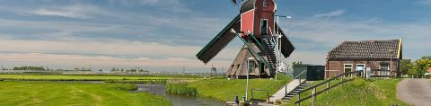 Eine Windmühle vor einem kleineren Haus. Dahinter weites, flaches Grasland in Südholland.