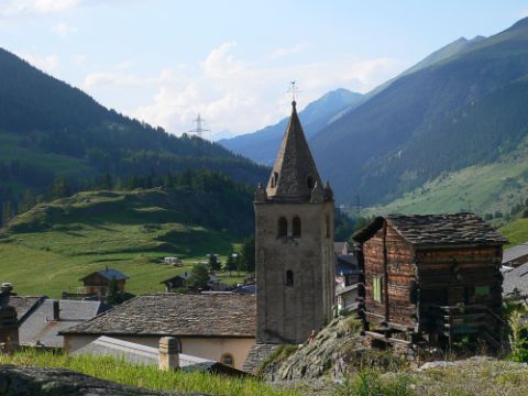 Rechts hat es ein kleines Holzhüttchen und gleich daneben der Glockenturm einer alten Kirche. Im Hintergrund ein Bergpanorama mit kleinen Wolken.