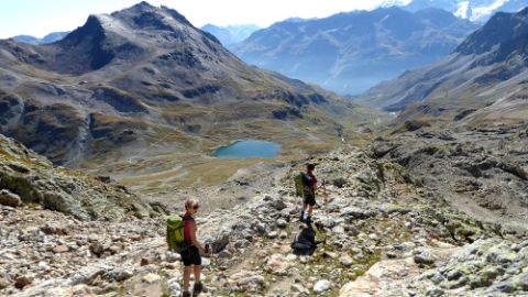 2 Wanderinnen laufen dem kleinen Bergsee entgegen mit Bergen im Hintergrun.