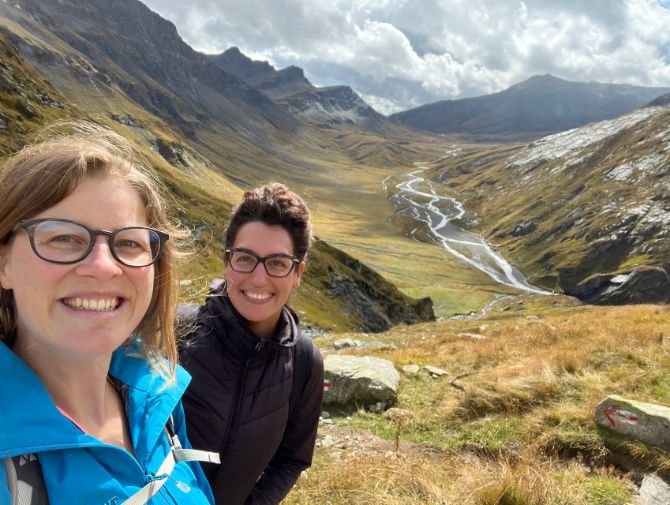 Zwei Frauen grinsen in die Kamera, im Hintergrund die Greina auf dem Alpenpässeweg zu sehen. 