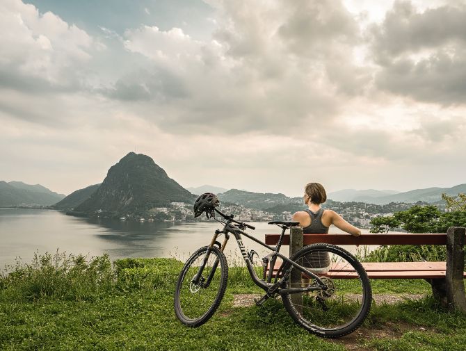 Eine Frau sitzt auf einer Bank mit Sicht auf den Lago di Lugano und den San Salvatore.