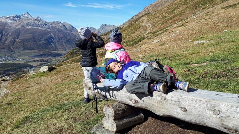 Drei Kinder machen Pause. Ein Mädchen liegt auf einem Holzbank, die anderen zwei schauen sich die Berge an.