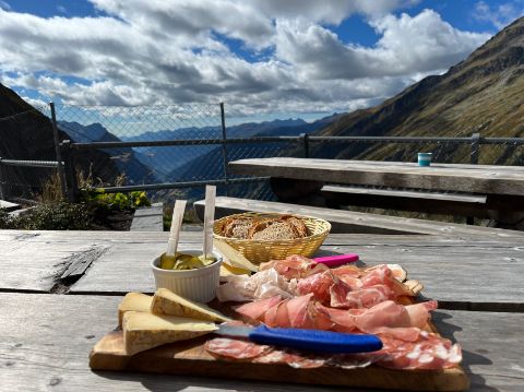 Ein Schinken-Plättchen und Käse auf einem Holztisch auf der Greina-Hochebene mit Aussicht ins Tal. 