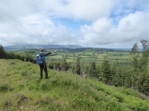 Auf dem grasigen Hügel steht ein Wanderer, der in die weite, grüne Landschaft zeigt, die tiefer unten ist. Der Himmel ist hellblau aber mit vielen Cumulus Wolken bedeckt.