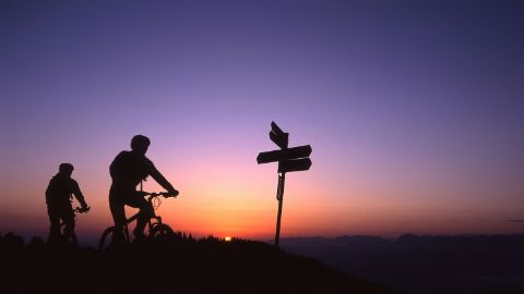 Zwei Mountainbiker radeln bei Sonnenuntergang an einem Wegweiser vorbei.