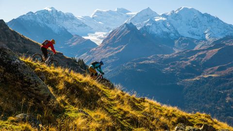Zwei Trailrunner fahren auf einem Naturweg den Berg hinunter. Im Hintergrund eine zauberhafte Berglandschaft. Teilweise sind die Bergspitzen mit Schnee bedeckt.