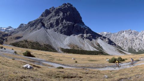 Deux vététistes passent devant les montagnes grisonnes, sur un étroit chemin de pierres naturelles.