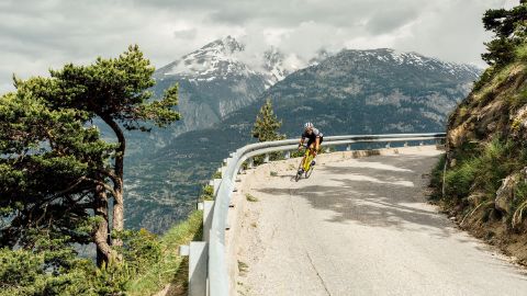 Cycliste sortant d'un virage dans un paysage de montagne avec des sommets enneigés.