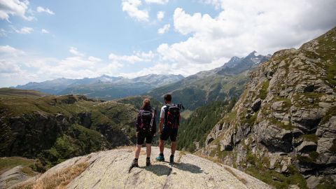 A couple of hikers stand on a stone ledge and look at the surrounding mountains.