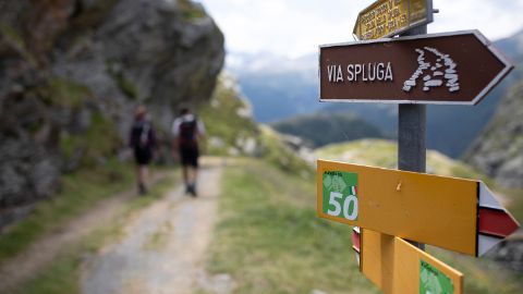 A signpost for the path towards Via Spluge in front of a blurred picture of 2 hikers on a natural road.