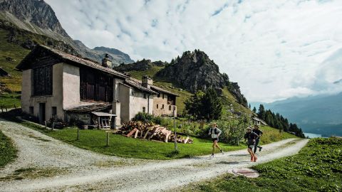 Jogger rennen an einem alten Steinhaus vorbei in den Bergen. Im Hintergrund sieht man den See. 