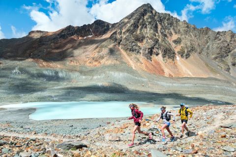 Three trail runners jog along a narrow natural stone path next to a mountain lake over the via Grischuna. In the background, the massive mountain under a slightly cloudy blue sky.